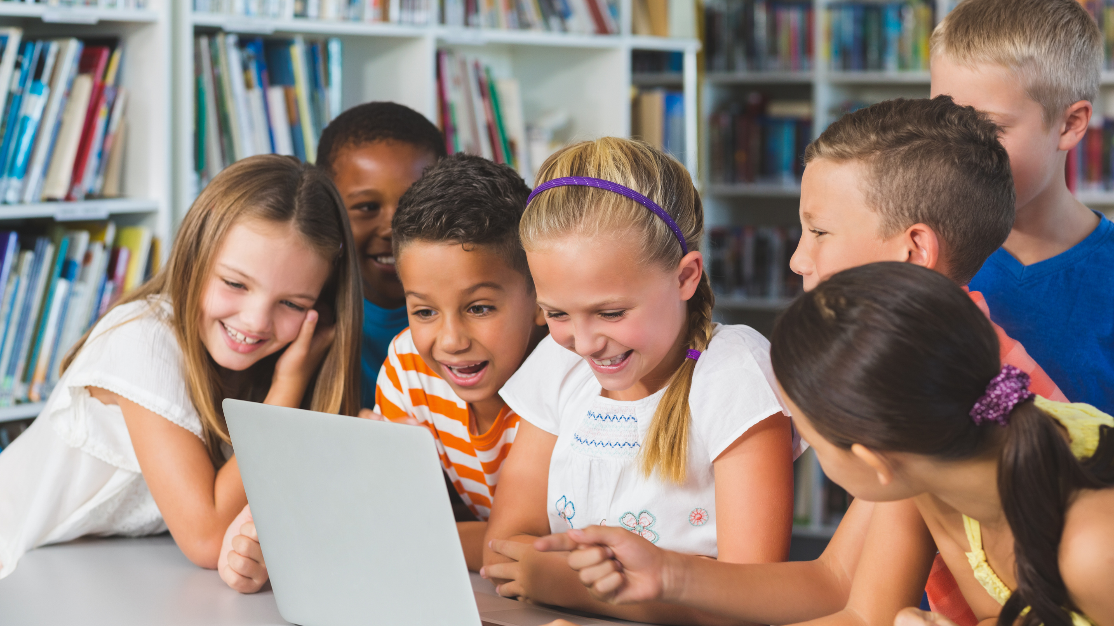 Kids of different backgrounds or ethnicities are gathered around a laptop smiling and laughing, in a library.