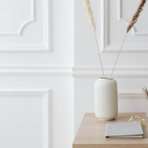 A white ceramic vase with dried grass stems placed on a wooden table beside a closed notebook